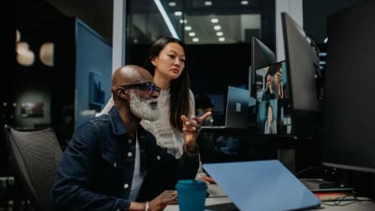 Two professionals working at a desk with a video call on a monitor