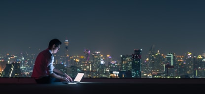 A person using a laptop on a rooftop at night with a city skyline in the background